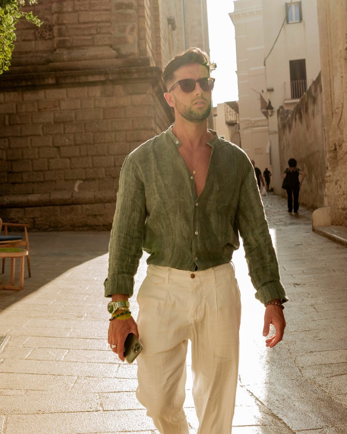 Man in green linen shirt and white trousers walking through a sunlit narrow street.