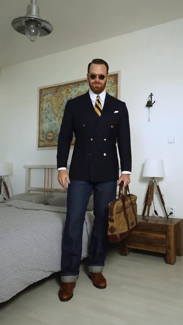 Man wearing a navy double-breasted blazer, striped tie, and dark jeans standing in a bedroom.