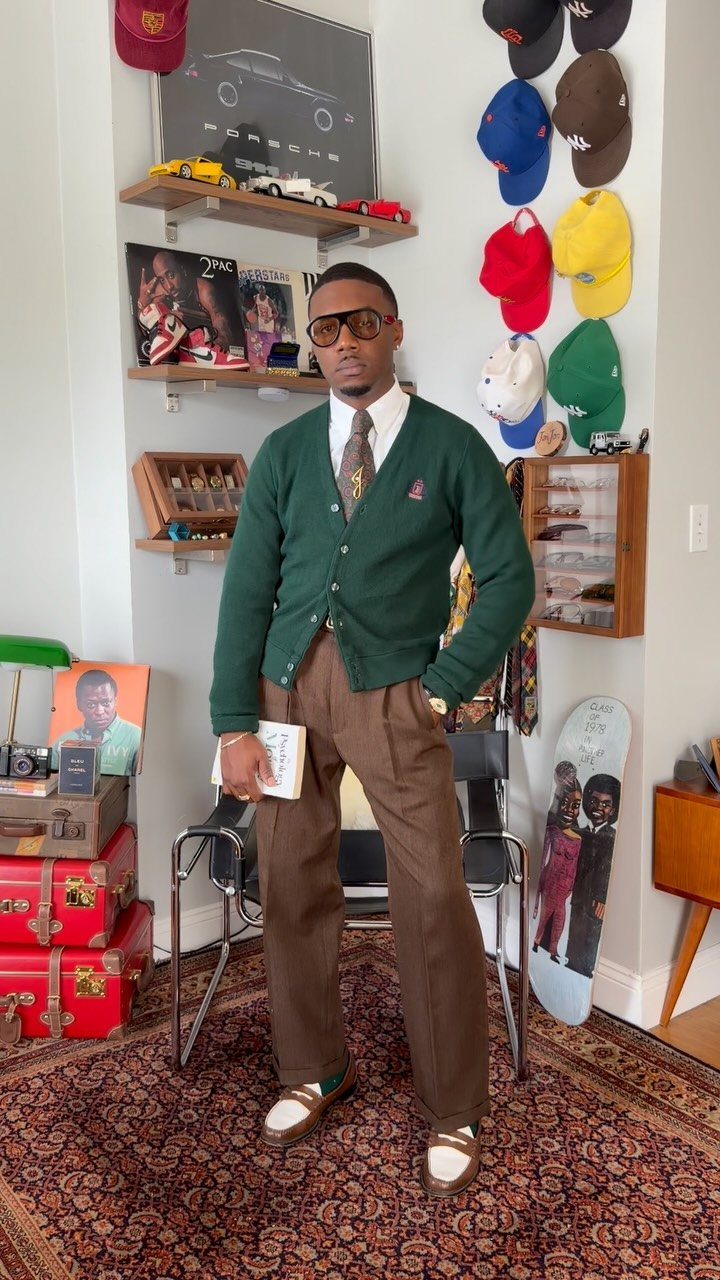 A man wearing a green cardigan, brown tie, and brown trousers posing in a room.