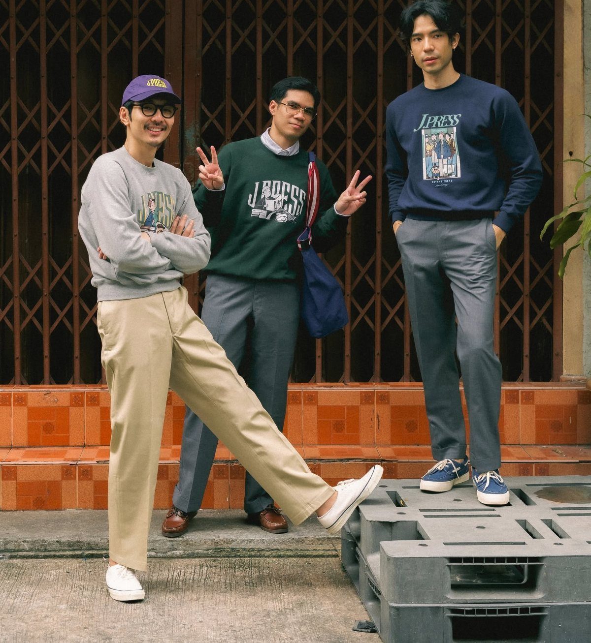 Three men posing in casual sweatshirts and trousers against a dark patterned wall.