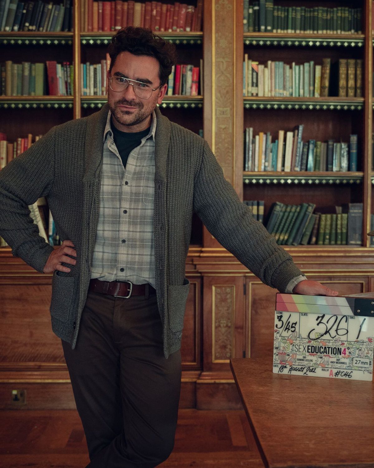 A man wearing a grey knit cardigan and plaid shirt standing in a library.