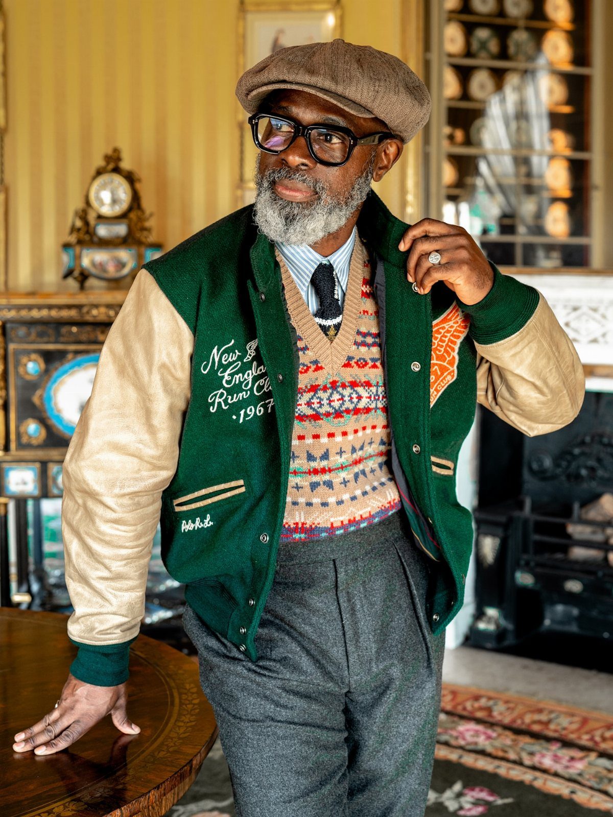 Man wearing a tan and green letterman jacket, patterned sweater vest, and a brown flat cap.