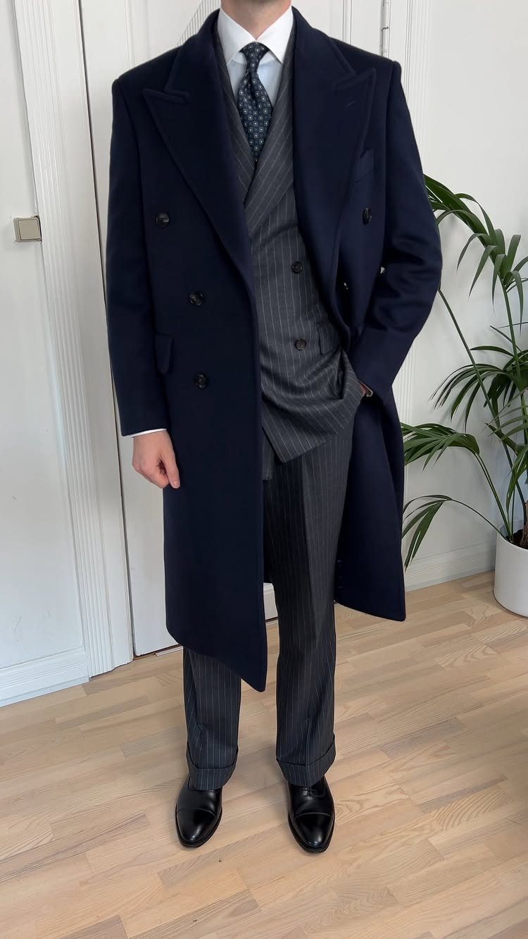 A person dressed in a navy overcoat, grey pinstripe suit, and black leather shoes.