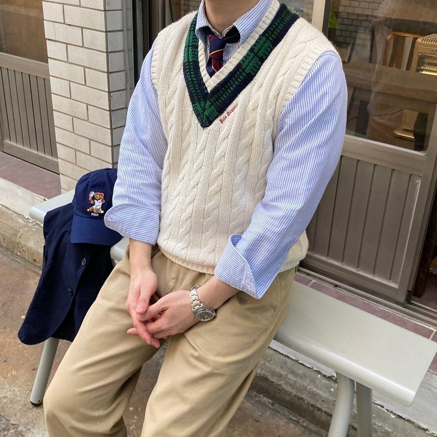 A person wearing a cream cable-knit sweater vest with plaid trim over a striped shirt and tie.