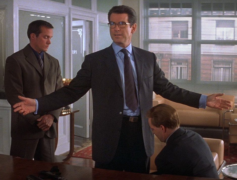 Man in a dark charcoal tailored suit and blue shirt standing with arms outstretched in an office.