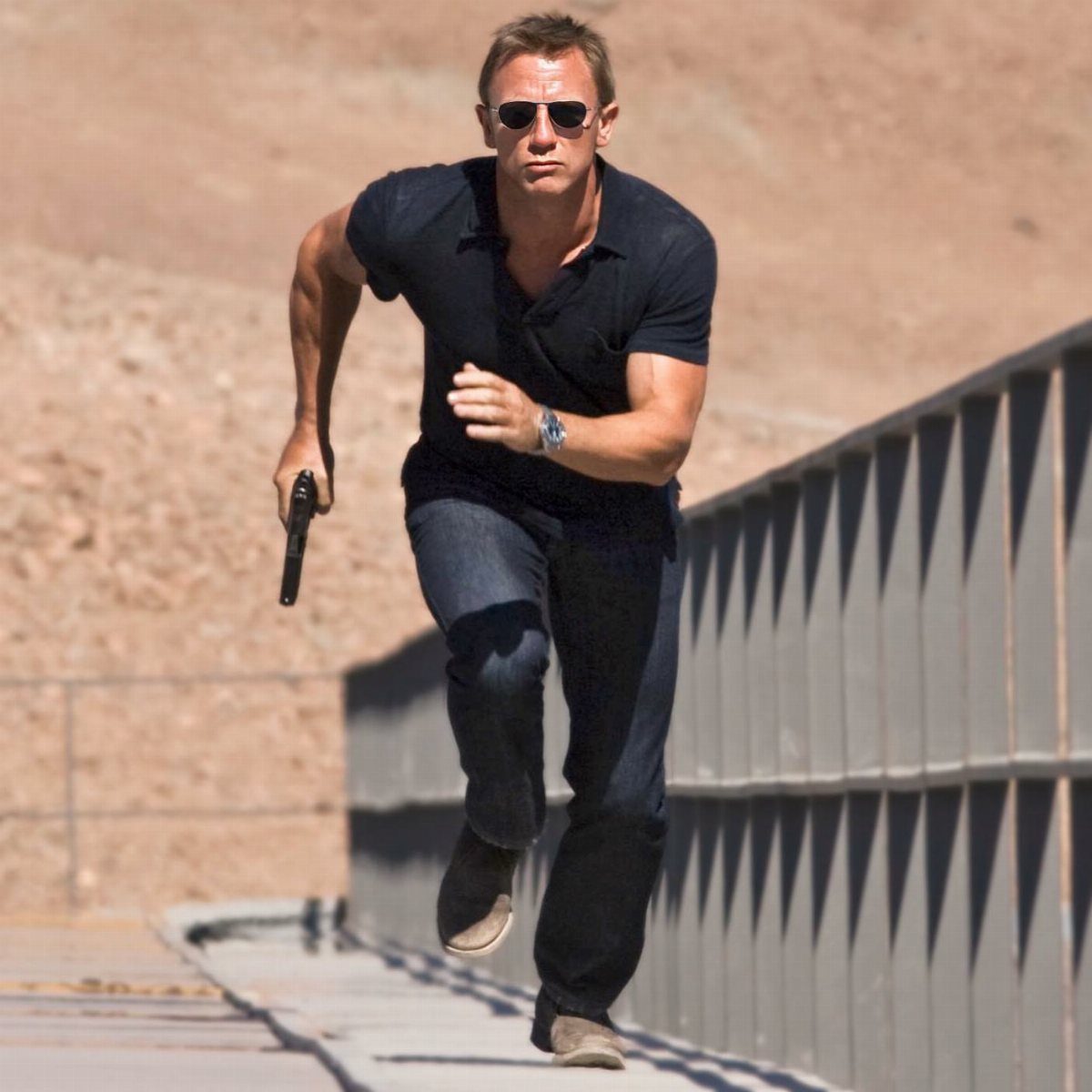 A man in sunglasses and a dark polo shirt runs while holding a handgun.