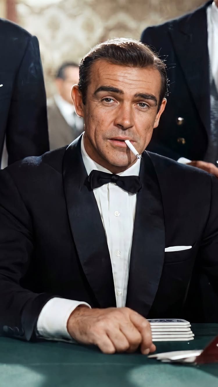 Man in a black tuxedo with a cigarette in his mouth sitting at a green gambling table.