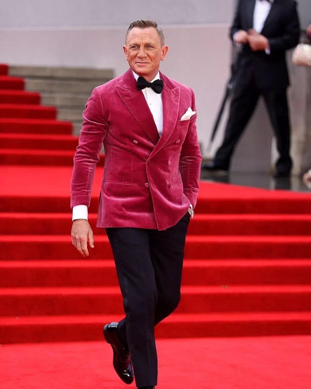Daniel Craig walking on a red carpet wearing a pink velvet dinner jacket and black bow tie.