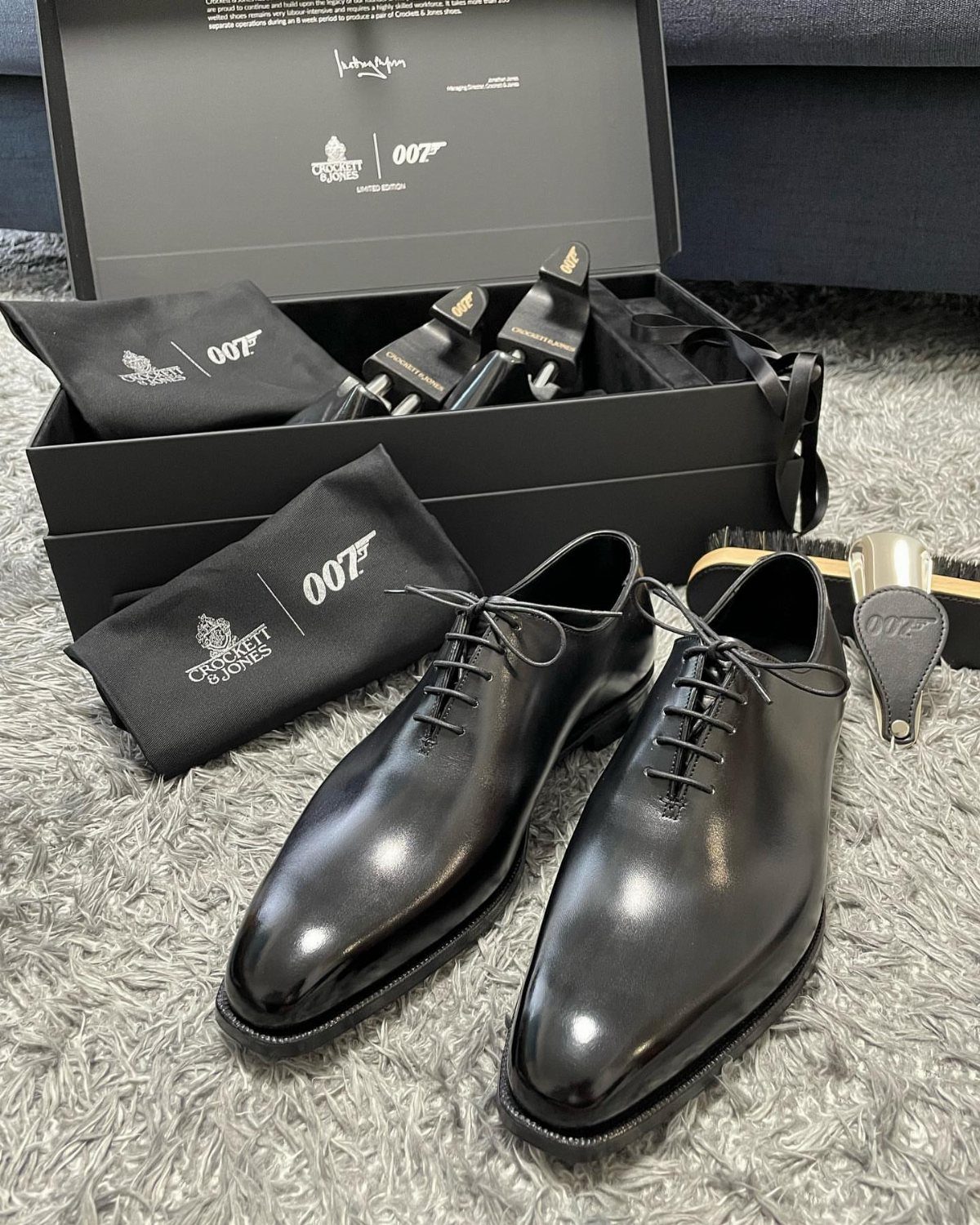 Polished black leather derby shoes displayed with a 007 branded gift box on a grey rug.