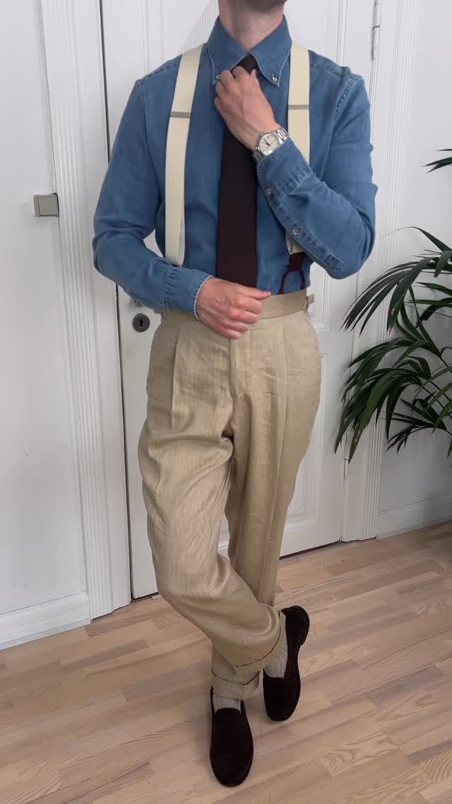 Man wearing a blue shirt, brown tie, cream suspenders, and beige trousers with dark loafers.
