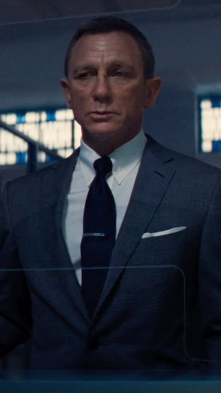 Daniel Craig wearing a dark grey tailored suit and a crisp white dress shirt with a navy tie.