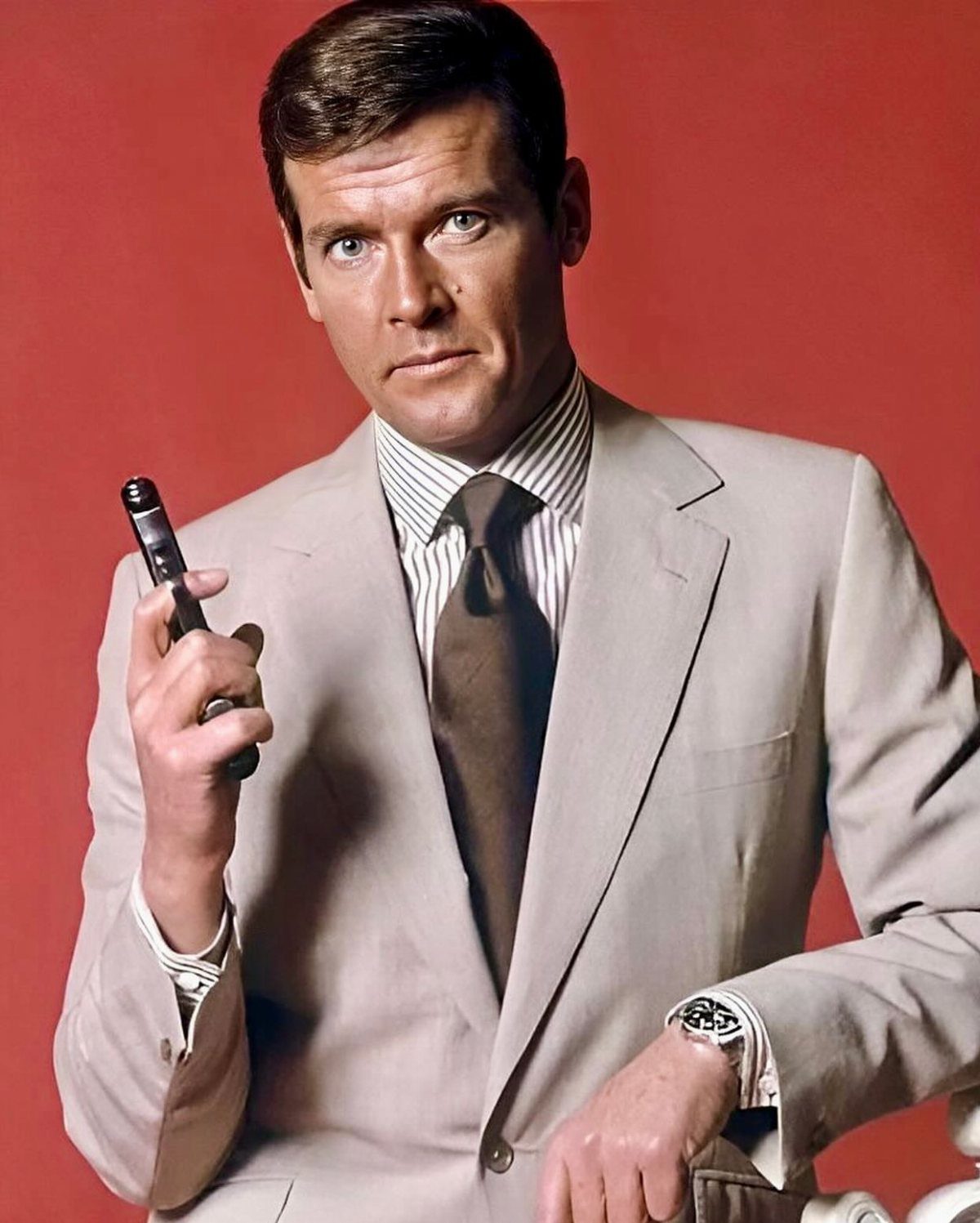 Man in a tan suit, striped shirt, and brown tie posing against a red background.