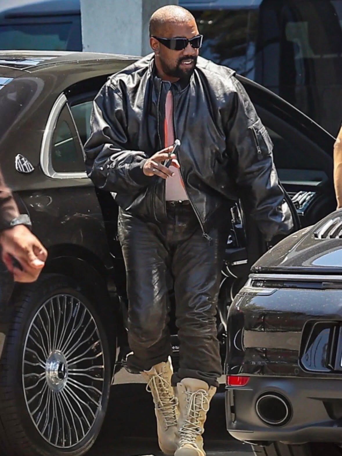Kanye West wearing a black leather jacket and pants while exiting a dark car outdoors.