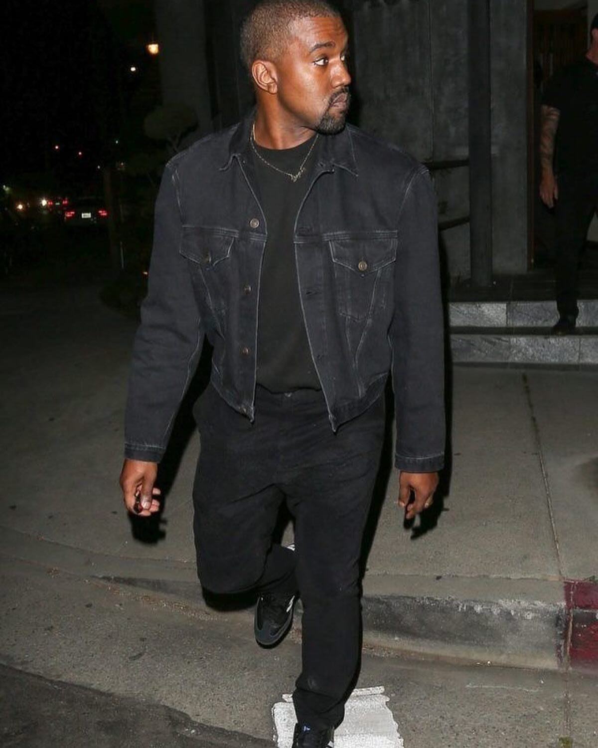Kanye West wearing a black denim jacket, black t-shirt, and black jeans while walking at night.