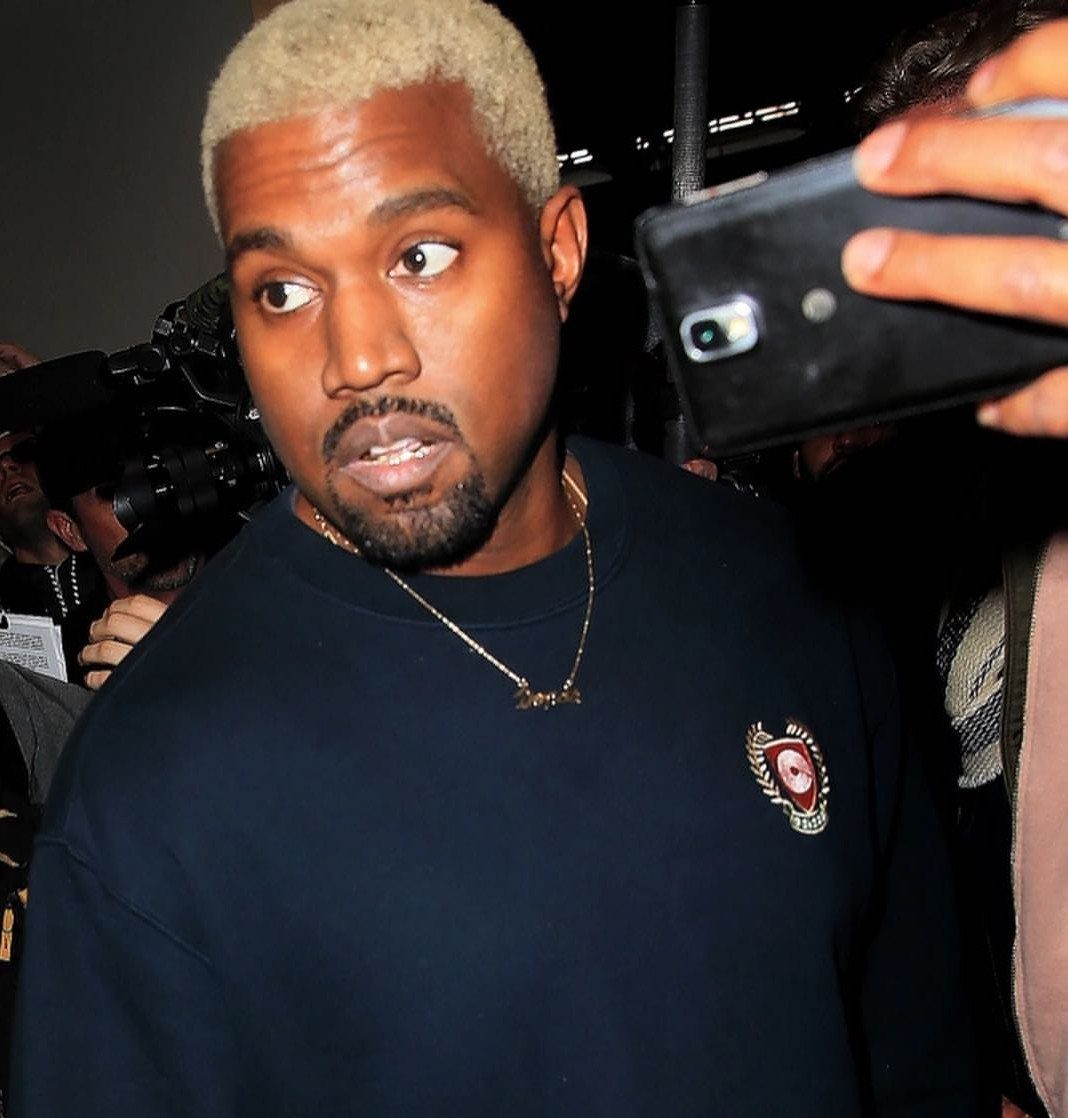 Kanye West with short bleached blonde hair looking sideways in a crowded room.