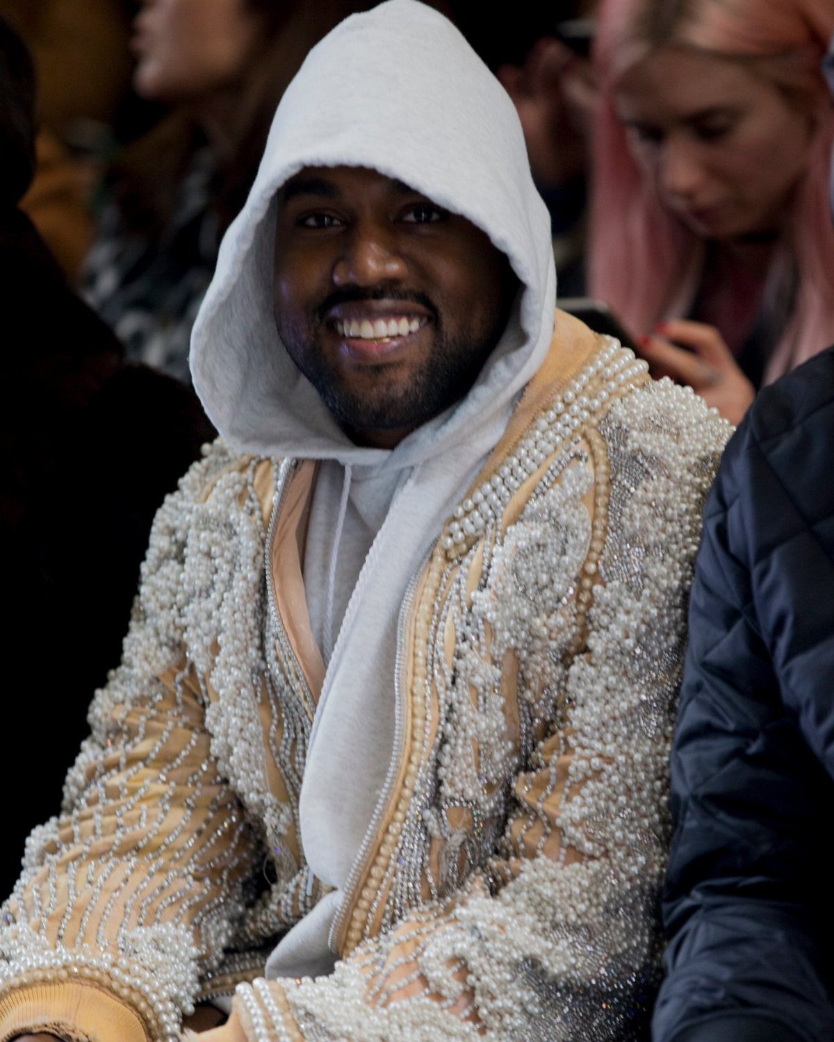 Kanye West smiling in a white hoodie and a pearl-encrusted beige jacket.