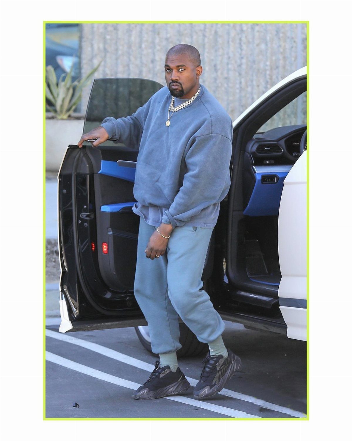 Kanye West posing by a car door wearing a grey sweatshirt and light blue sweatpants.
