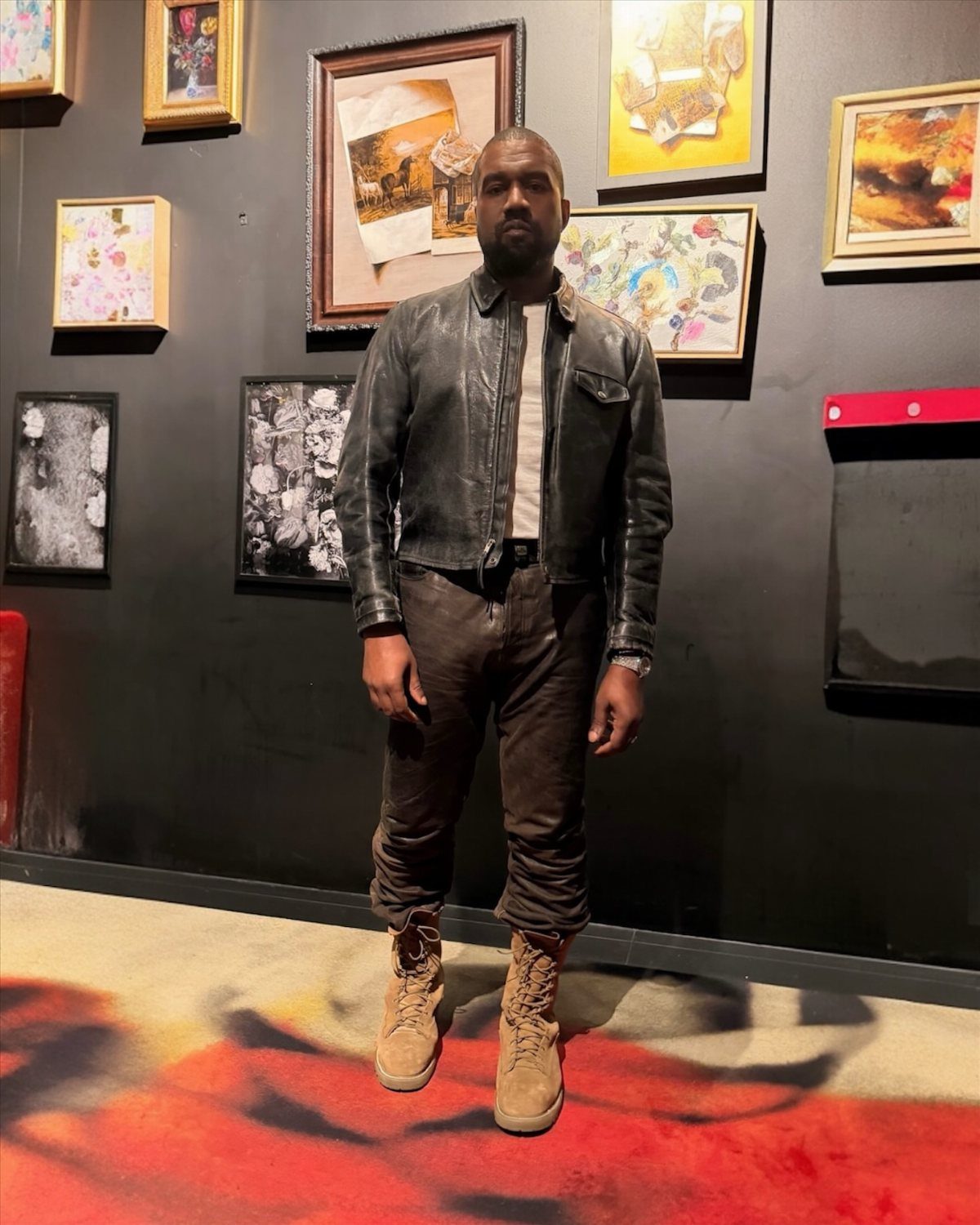 A man in a leather jacket, dark trousers, and tan boots stands before framed art.