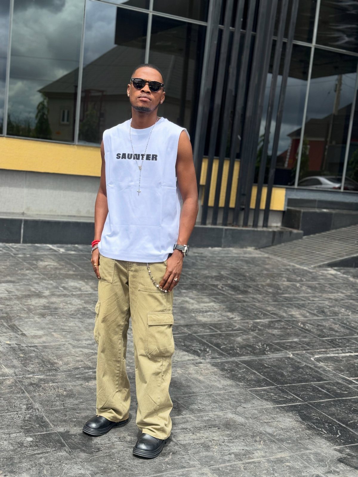 Man wearing a white sleeveless shirt, khaki cargo pants, and black sunglasses standing outdoors.