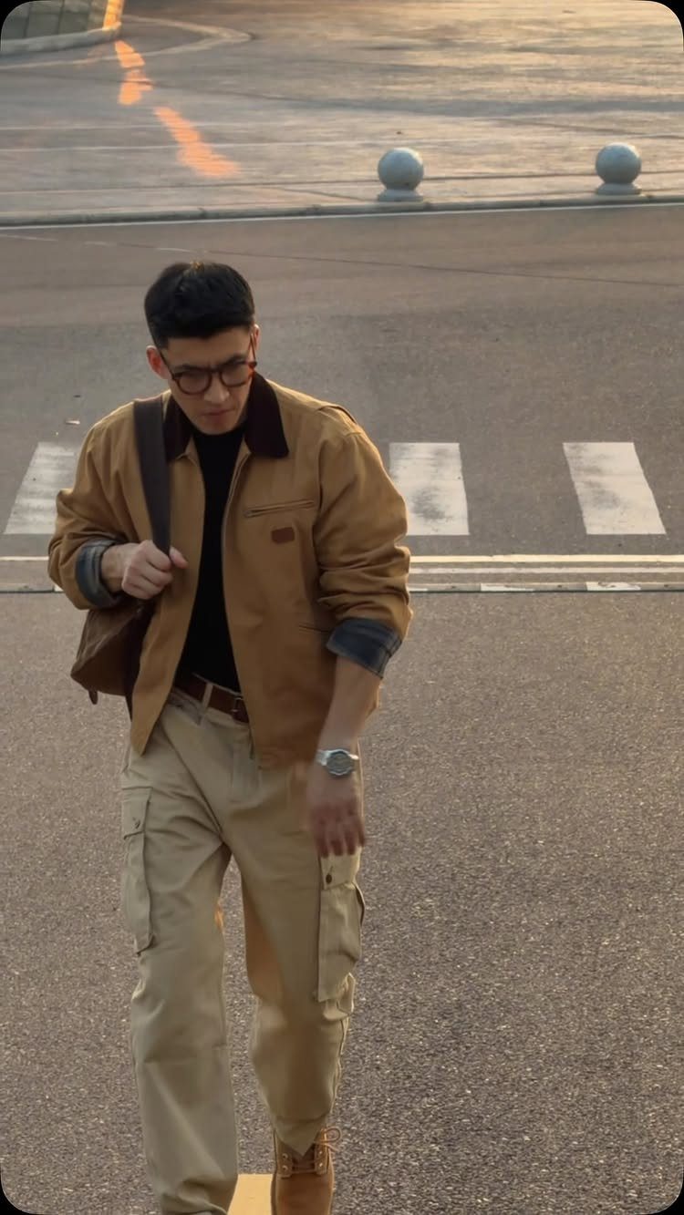A man in a tan utility jacket and khaki cargo pants walking across a street.