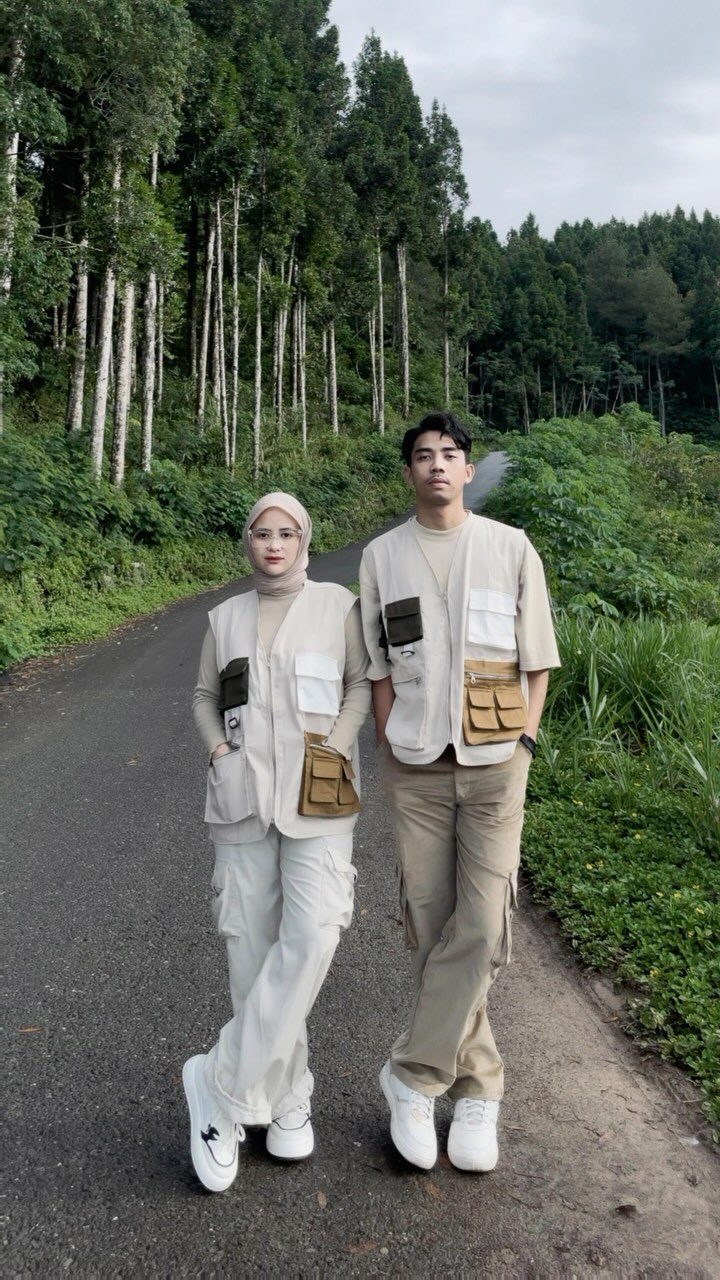 A man and a woman posing in khaki cargo pants and utility vests on a forest road.