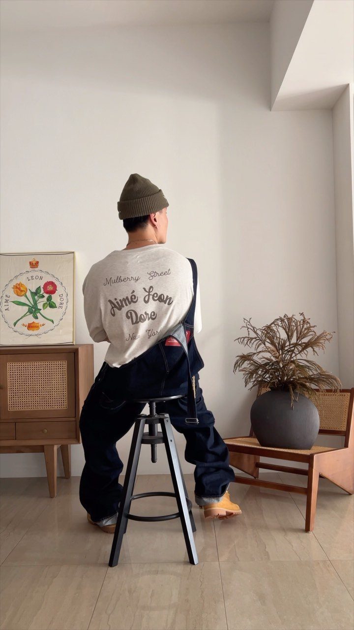 A person wearing a white graphic t-shirt and dark overalls sitting on a stool.