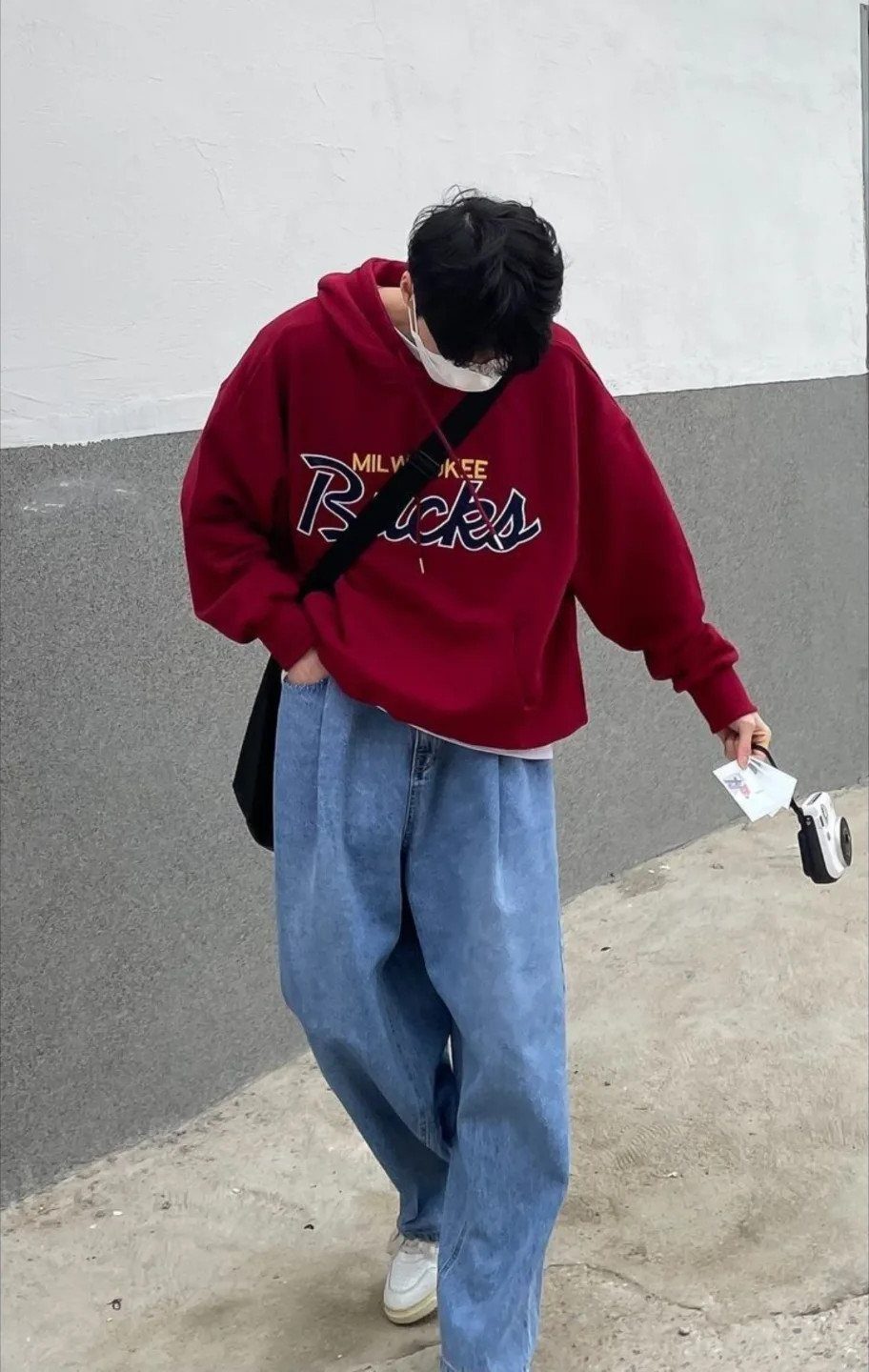 A person walking in a red hoodie and baggy blue jeans against a neutral wall.