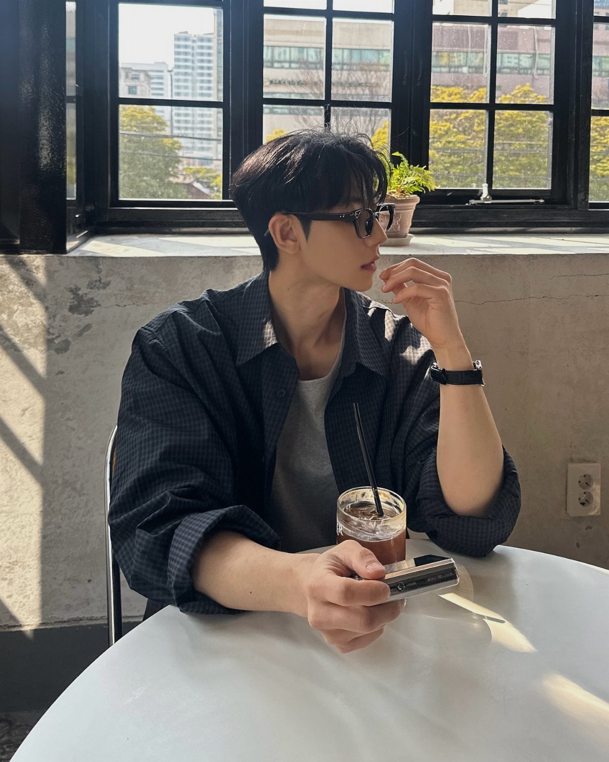 A young man wearing a dark button-down shirt and glasses sits at a cafe table.