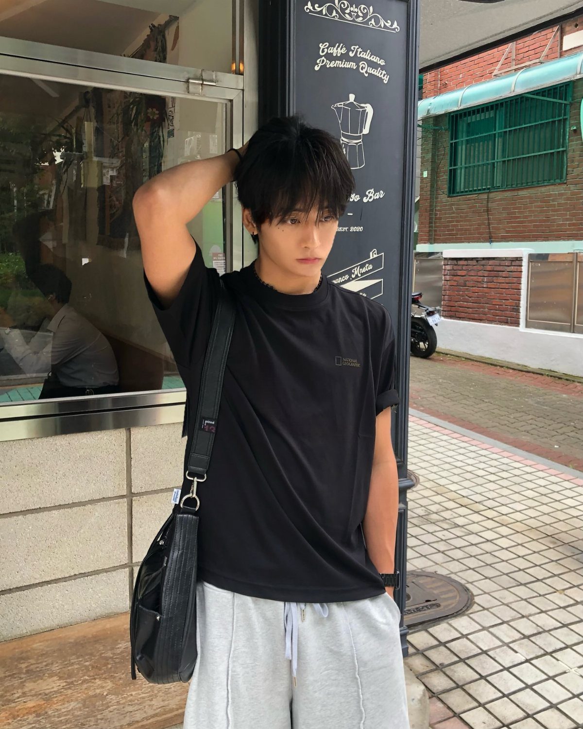 A young man in a black t-shirt and grey sweatpants standing by a shop.