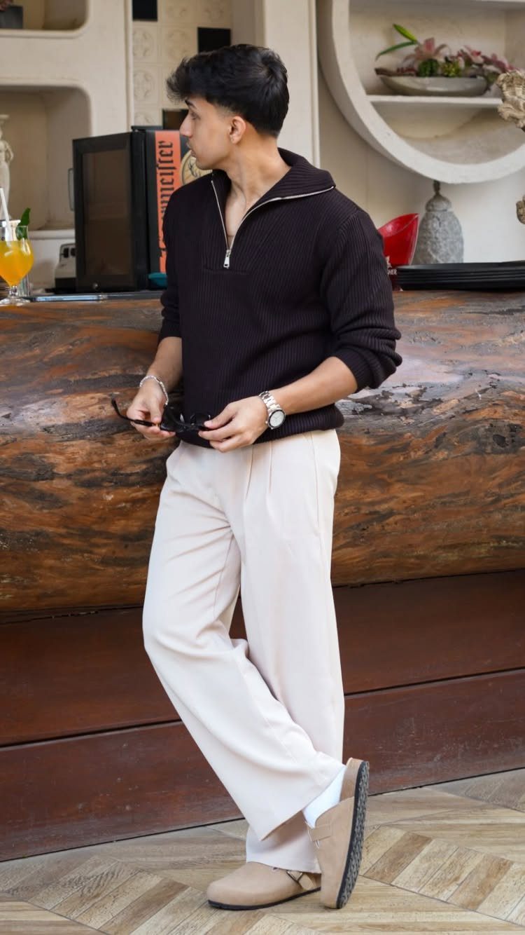 A man wearing a black quarter-zip sweater and cream wide-leg trousers standing by a wooden bar.