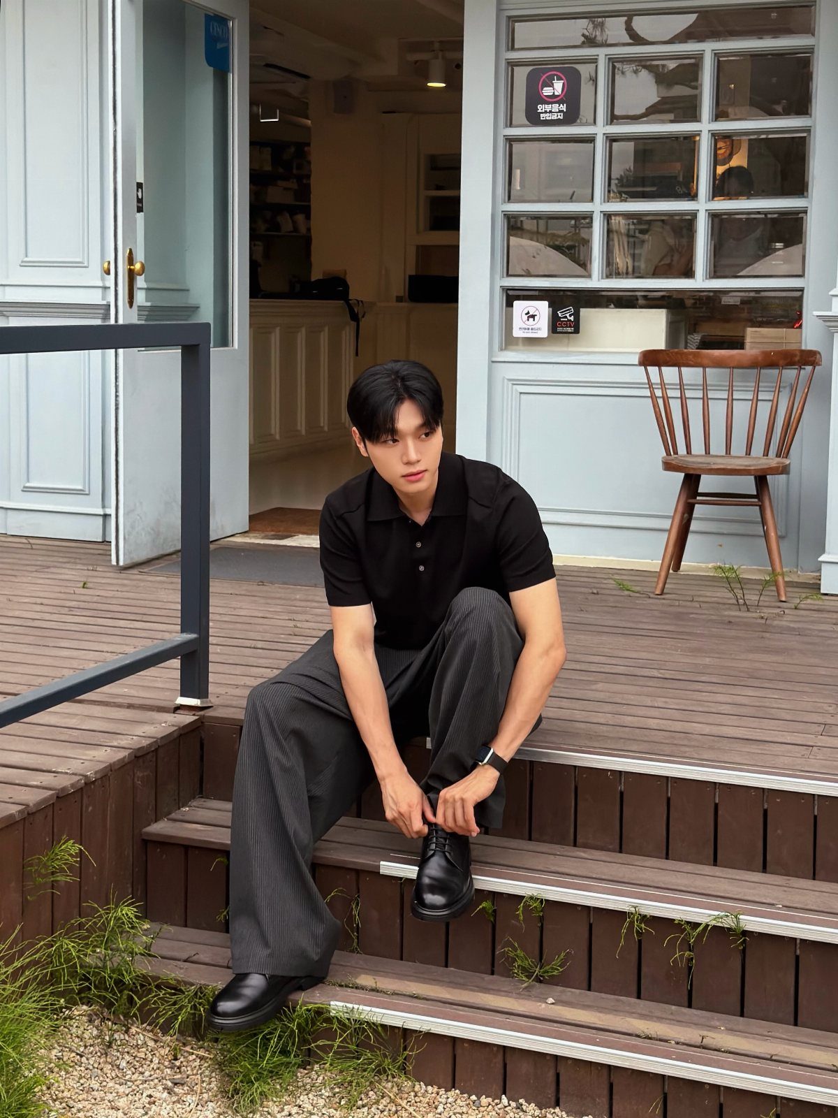 Man in a black polo and grey pinstriped trousers sitting on wooden outdoor steps.