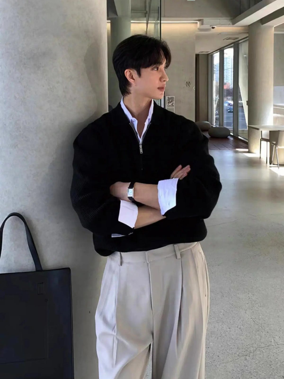 A young man in a black pullover and beige trousers leaning against a pillar.