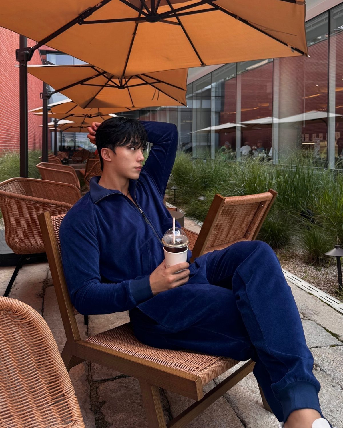 A young man in a navy blue velour tracksuit sitting at an outdoor cafe under orange umbrellas.