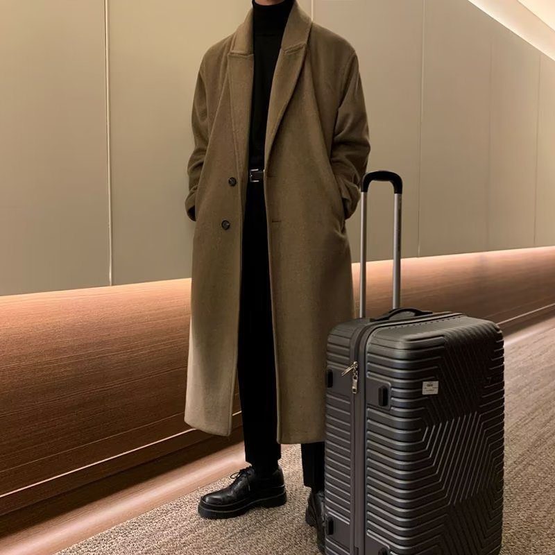 A person wearing a long tan wool coat and black turtleneck standing by a suitcase.