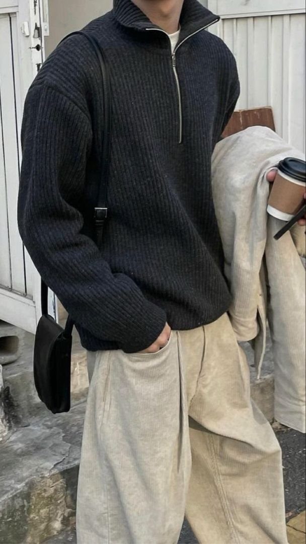 Person wearing a dark ribbed half-zip sweater and wide-leg beige trousers holding a coffee cup.