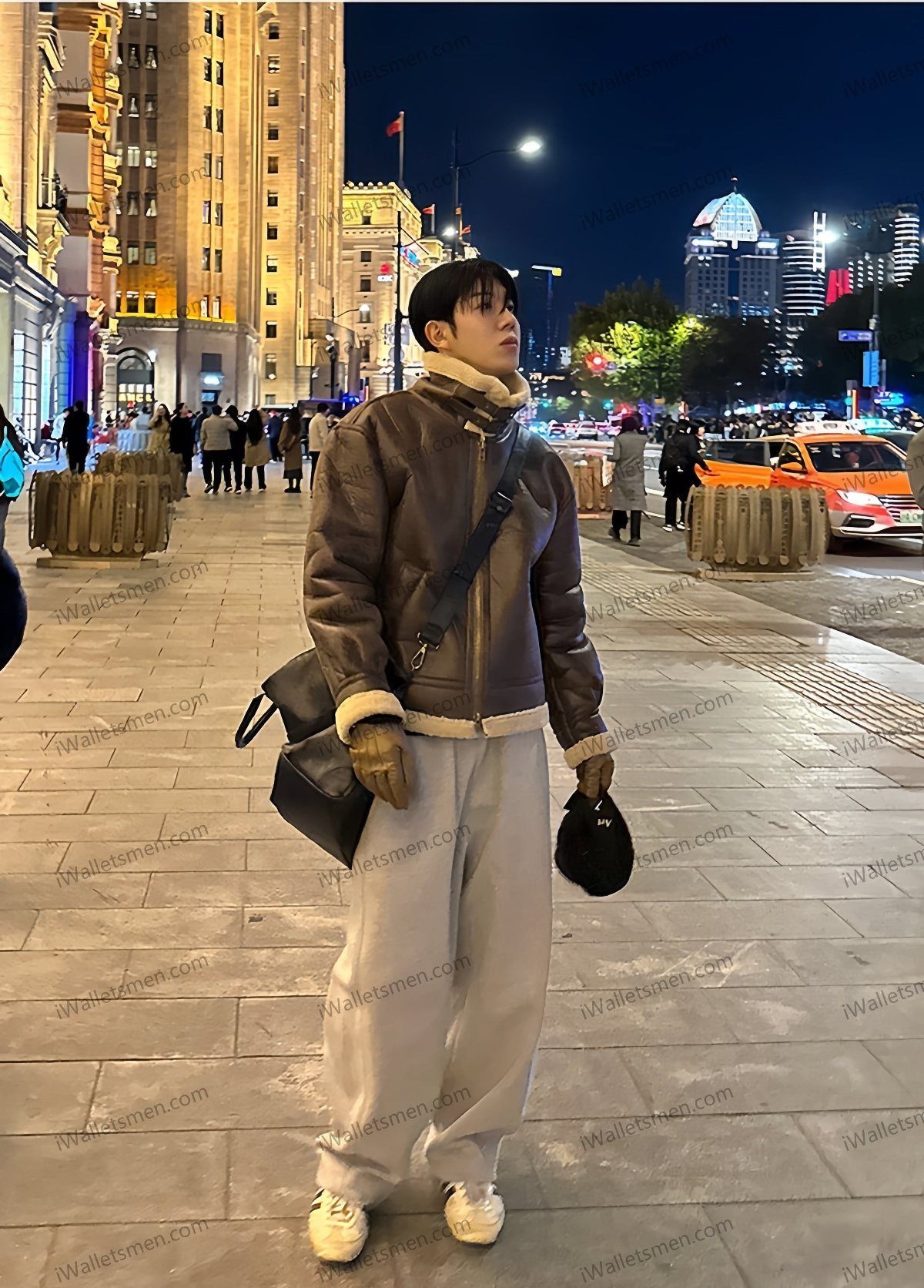 A person wearing a brown shearling jacket and wide-leg grey pants standing on a city street at night.