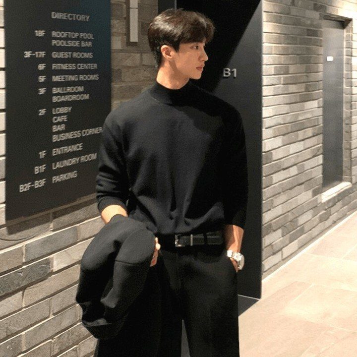 A man wearing a black slim-fit turtleneck sweater and black trousers standing by a brick wall.