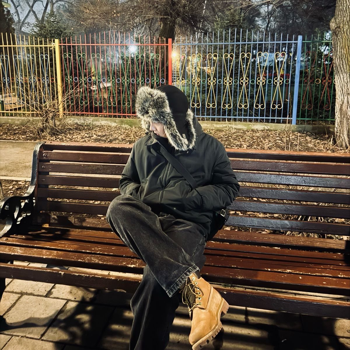 A person wearing a black jacket and a fur-trimmed ushanka hat sitting on a park bench.