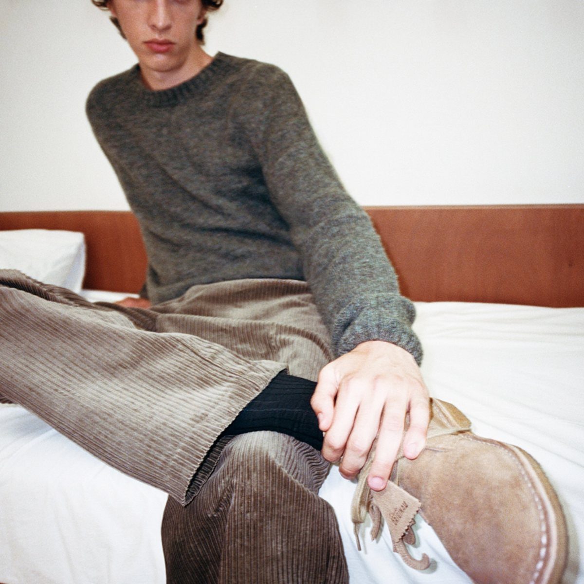 A man sitting on a bed wearing a grey sweater, corduroy pants, and suede boots.