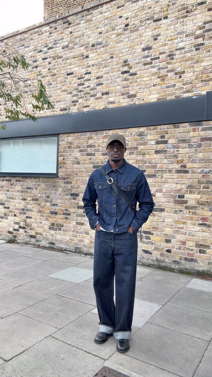 A man wearing a dark denim jacket and matching denim jeans standing against a brick wall.