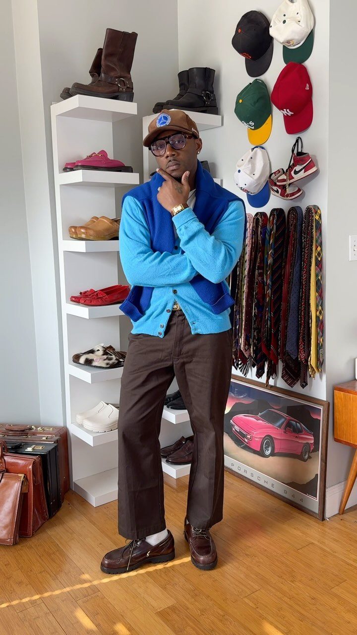 A man wearing a bright blue cardigan, blue sweater vest, brown trousers, and a brown cap.