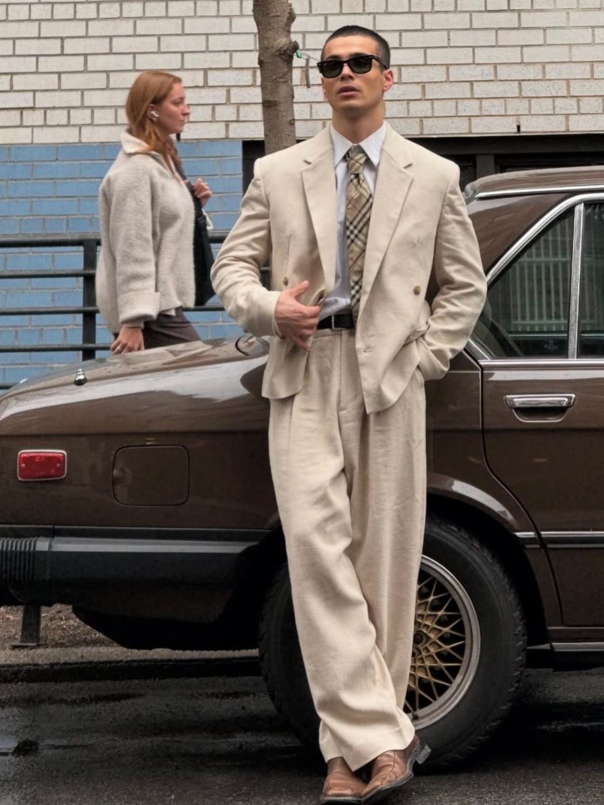 Man in a cream linen suit and patterned tie leaning against a brown vintage car.