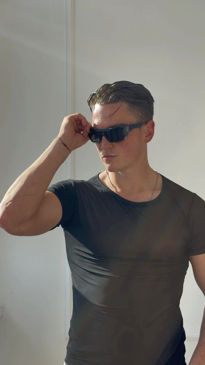 A young man in a sheer black t-shirt and sunglasses posing against a white wall.