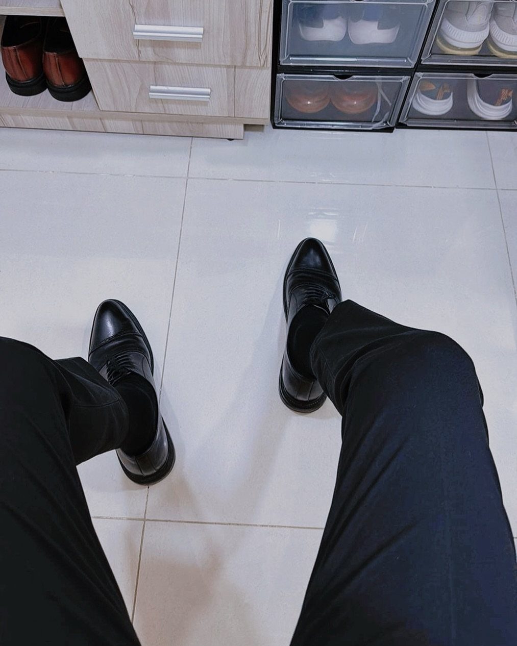 Black cap-toe oxford shoes worn with black trousers on a white tiled floor.
