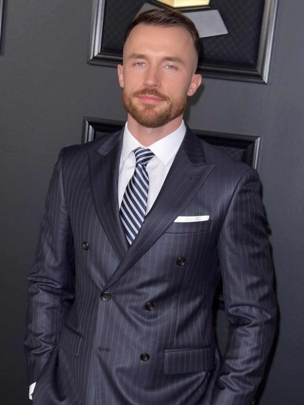 A man with groomed hair and a beard wearing a navy pinstripe suit and striped tie.