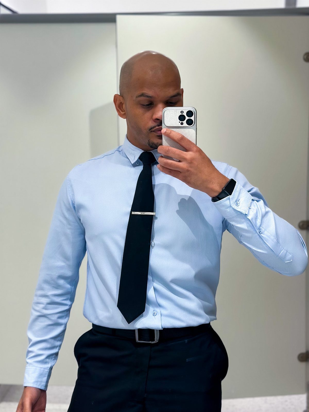 Man in a light blue shirt, black tie, and navy trousers taking a mirror selfie.