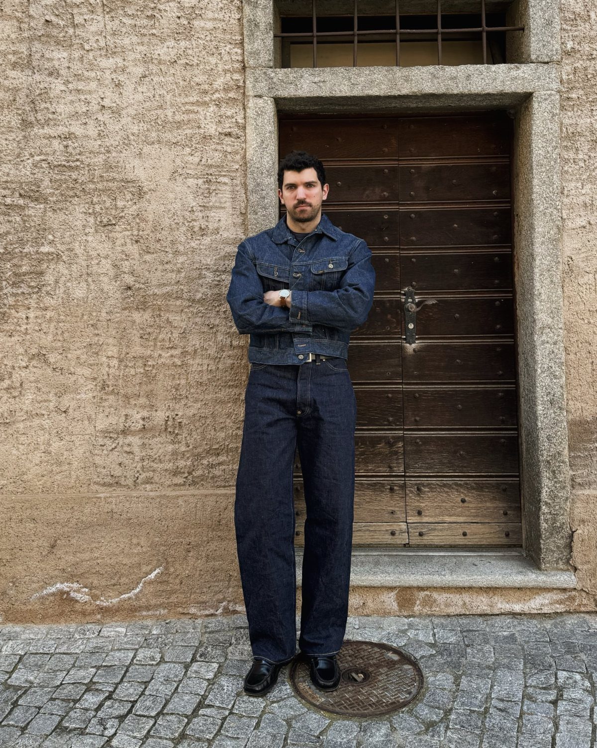 Man wearing a dark denim jacket and wide-leg denim jeans standing before a wooden door.