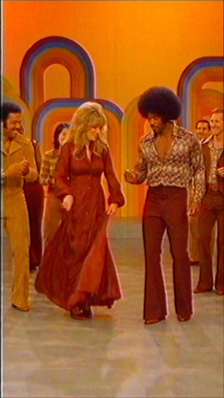 Man with an afro in patterned shirt and flared trousers walking on a stage.