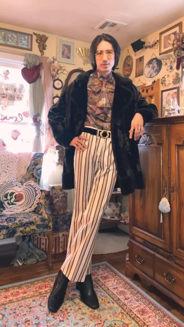 Man in a black fur coat, patterned shirt, and striped trousers posing indoors.