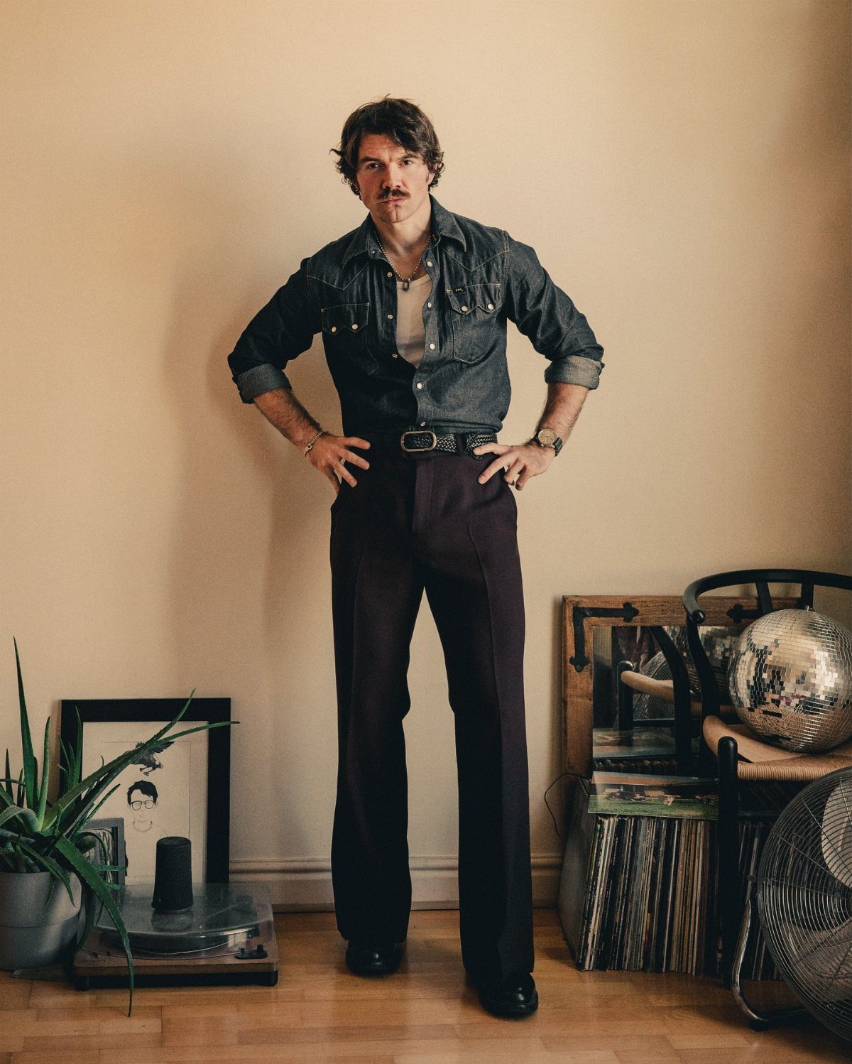 Man posing in a denim shirt and dark brown flared trousers against a neutral wall.